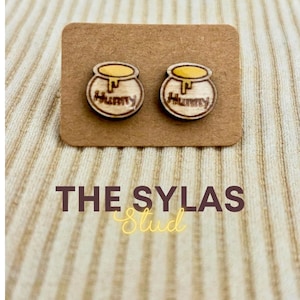 May include: Wooden stud earrings featuring a honey pot design with the word "Hunny" on each. The earrings are displayed on a brown card with the text "THE SYLAS Stud" below.