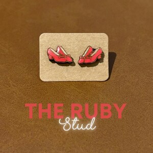 May include: A pair of red high-heeled shoe stud earrings with gold accents, displayed on a small, rectangular, tan card. The text "THE RUBY Stud" is below the earrings in red and white.