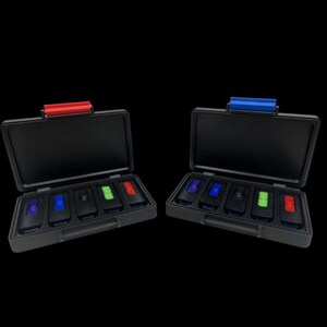 May include: Two black rectangular storage cases with hinged lids, each containing five small black rectangular components with colored tops. The left case features a red latch, while the right case features a blue latch. The components have purple, blue, black, green, and red accents.