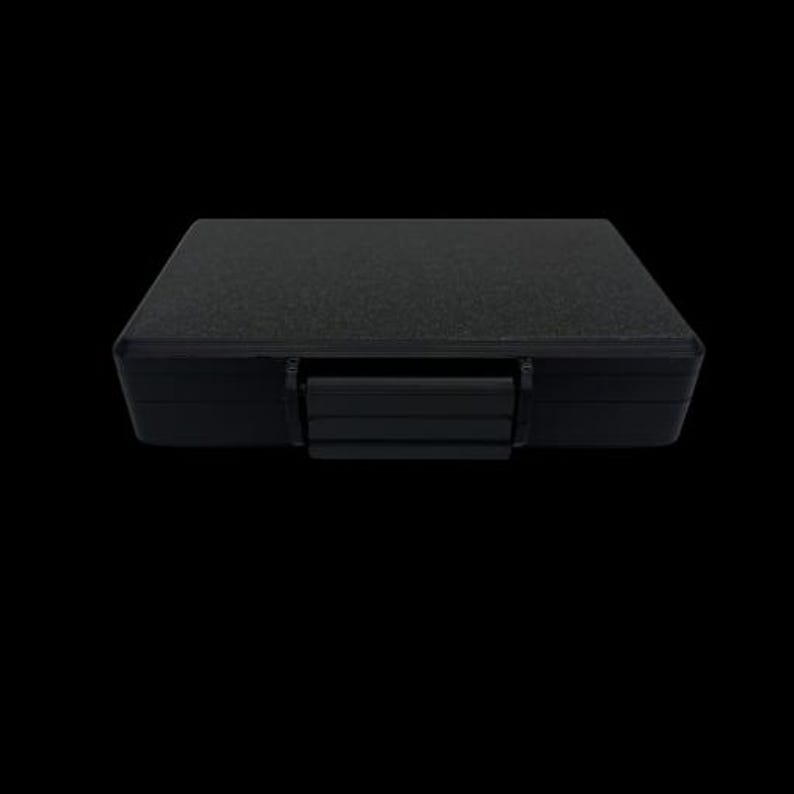 May include: A rectangular black storage case with a textured surface and a central latch mechanism. The case features a solid, opaque construction with a flat top and a secure locking clip positioned on the front edge for closure and protection.
