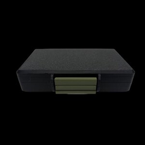 May include: A rectangular black storage case with a textured top surface and a solid olive green plastic latch centered on the front edge. The case features a sleek, low-profile design with rounded corners and a matte finish on the main body.