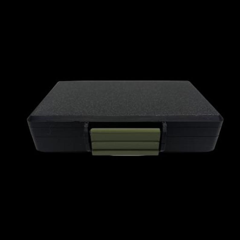 May include: A rectangular black storage case with a textured top surface and a solid olive green plastic latch centered on the front edge. The case features a sleek, low-profile design with rounded corners and a matte finish on the main body.