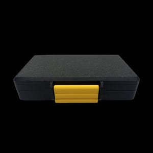 May include: A rectangular black storage case with a textured surface and a single bright yellow plastic latch centered on the front edge. The case features a low-profile design with rounded corners and a solid, opaque construction for secure storage.