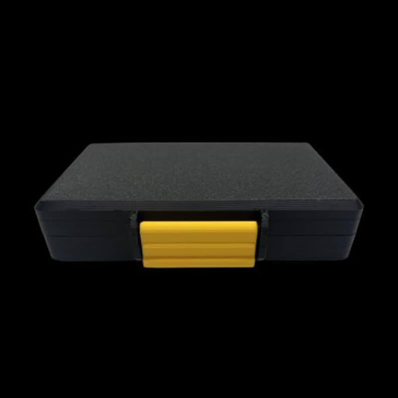 May include: A rectangular black storage case with a textured surface and a single bright yellow plastic latch centered on the front edge. The case features a low-profile design with rounded corners and a solid, opaque construction for secure storage.