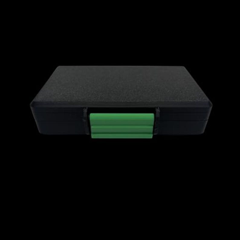 May include: A rectangular black storage case with a textured surface and a bright green plastic latch centered on the front edge. The case features a solid, opaque construction with a flat top and a secure locking mechanism for closure.