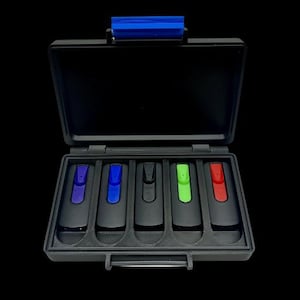 May include: A black rectangular storage case with a blue latch containing five individual slots. Each slot holds a black electronic device featuring a distinct colored clip in purple, blue, black, green, and red, arranged in a row inside the case.