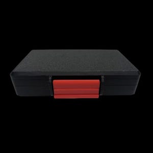 May include: A rectangular black storage case with a textured top surface and a single bright red plastic latch centered on the front edge. The case features a solid black frame and a flat, matte black lid, providing a secure and simple container.