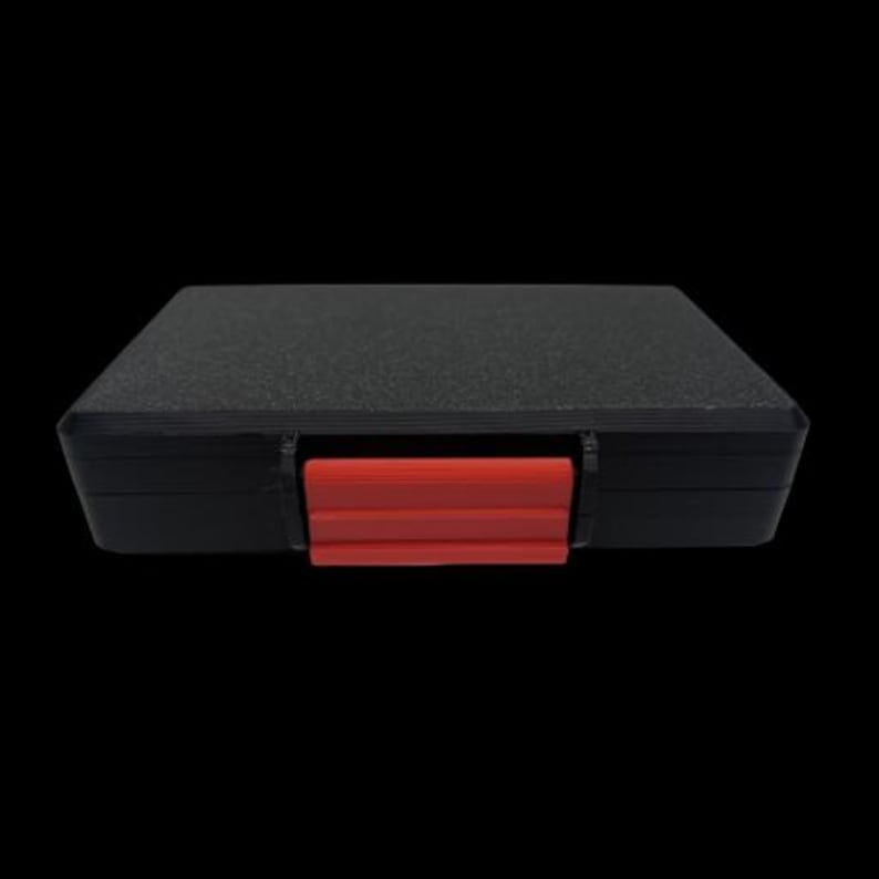 May include: A rectangular black storage case with a textured top surface and a single bright red plastic latch centered on the front edge. The case features a solid black frame and a flat, matte black lid, providing a secure and simple container.