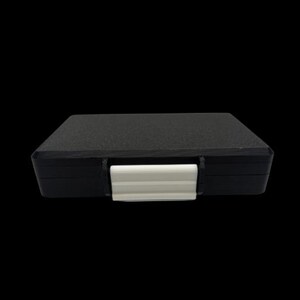 May include: A rectangular black storage case with a textured surface and a smooth white plastic latch centered on the front edge. The case features a flat top and a solid construction, designed for secure closure and protection of its contents.