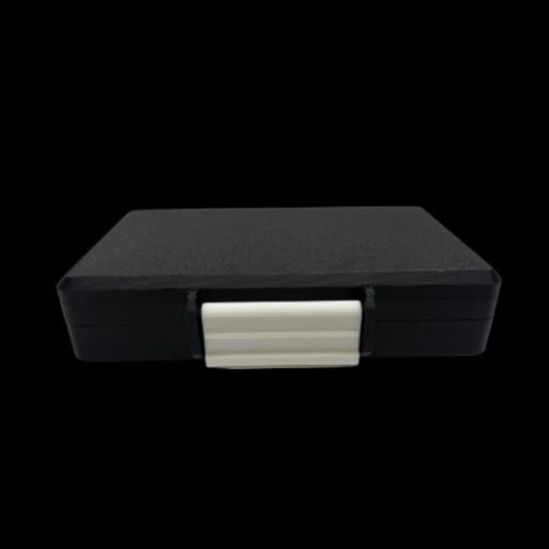 May include: A rectangular black storage case with a textured surface and a smooth white plastic latch centered on the front edge. The case features a flat top and a solid construction, designed for secure closure and protection of its contents.