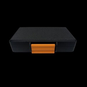 May include: A rectangular black storage case with a textured surface and a single bright orange plastic latch centered on the front edge. The case features a solid, opaque construction with clean, straight lines and a secure locking mechanism.