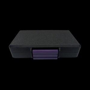 May include: A rectangular black storage case with a textured top surface and a solid purple latch centered on the front edge. The case features a smooth black frame and a secure closing mechanism, providing a simple and functional design for storage.