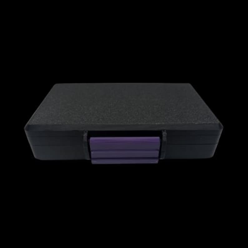 May include: A rectangular black storage case with a textured top surface and a solid purple latch centered on the front edge. The case features a smooth black frame and a secure closing mechanism, providing a simple and functional design for storage.
