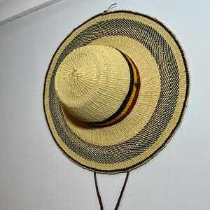 May include: A woven straw hat with a wide brim and a rounded crown. The hat features alternating bands of natural straw and black, with a decorative band of red, yellow, and black. Brown leather cords hang from the hat.
