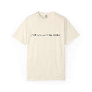 May include: Cream-colored t-shirt with the phrase "Plot twists are my cardio" printed in black. The shirt has a crew neck and short sleeves. The fabric appears to be soft and comfortable.
