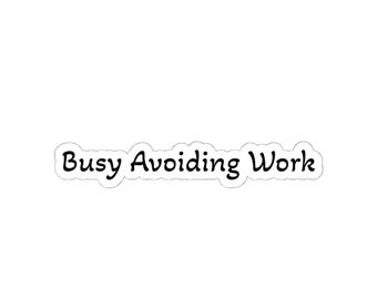 May include: A white sticker with the phrase "Busy Avoiding Work" in a black, handwritten-style font. The text is outlined in black and has a slight shadow effect, giving it a 3D appearance.