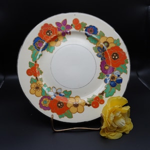 May include: A vintage cream-colored plate with a vibrant floral design in orange, red, yellow, purple, and blue. The flowers are arranged in a circular pattern around the plate's edge. A yellow flower rests near the plate.