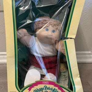 May include: A 1985 Cabbage Patch Kids doll with light brown yarn hair and blue eyes, wearing a white shirt and red pants. The doll is inside its original yellow and green cardboard box with a clear plastic window and includes an adoption certificate.