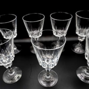 May include: Set of seven clear crystal wine glasses. Each glass features a stemmed base and a detailed, textured bowl. The glasses are arranged on a black surface, showcasing their elegant design and clarity.