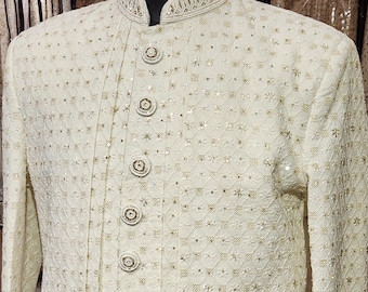 Può includere: Uno sherwani color bianco sporco con collo alto alla coreana e un'abbottonatura frontale con otto bottoni circolari decorati. Il capo è ricoperto da un fitto e complesso motivo di piccole paillettes argentate riflettenti e ricami chiari.