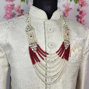 May include: A cream-colored, patterned jacket with a high collar and a decorative beaded necklace. The necklace features red and white beads, with a silver-colored clasp. The jacket is displayed on a mannequin.