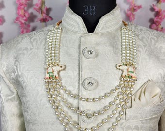 Può includere: Giacca color crema fantasia con collo alla coreana e bottoni decorativi. Una collana di perle a più fili con ciondoli a forma di elefante è drappeggiata sulla giacca. Un fazzoletto bianco è nella tasca sul petto.