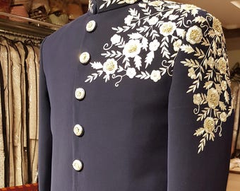 Puede incluir: Chaqueta formal azul marino con intrincados bordados florales blancos y dorados en los hombros y el cuello. La chaqueta presenta botones dorados grandes y redondos en la parte delantera y en los puños. La prenda se exhibe en un maniquí.