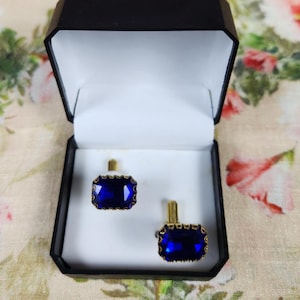 May include: A pair of cufflinks with rectangular, deep blue gemstone centers set in gold-toned metal. The cufflinks are displayed in an open black box with a white interior, set against a floral background.