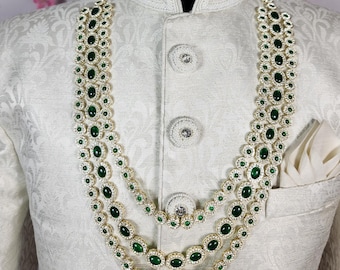 Puede incluir: Un collar de tres hilos con piedras verde esmeralda y detalles blancos se muestra sobre una chaqueta blanca. La chaqueta tiene un patrón texturizado y un pañuelo de bolsillo. El collar es en capas y ornamentado, con un diseño clásico.