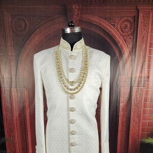 K&ouml;nnte beinhalten: Elfenbeinfarbene, lang&auml;rmlige formelle Jacke mit aufwendigen Details, auf einer Schaufensterpuppe pr&auml;sentiert. Die Jacke hat verzierte goldfarbene Kn&ouml;pfe und eine passende dekorative Halskette. Der Stoff hat ein subtiles, strukturiertes Muster, und der Kragen ist mit goldener Stickerei verziert.