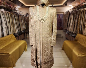 Puede incluir: Un traje sherwani dorado, bordado intrincadamente, exhibido en un maniquí. El conjunto incluye un turbante a juego con una pluma y un collar de perlas en capas. El traje se exhibe en una tienda de ropa.
