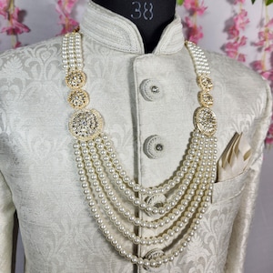 May include: A cream-colored, ornate necklace with multiple strands of faux pearls and gold-toned, circular embellishments. The necklace is displayed on a mannequin wearing a cream-colored, patterned jacket with a pocket square.