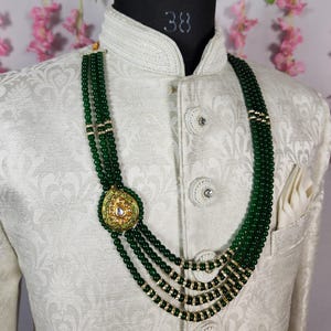 Puede incluir: Un collar de cuentas verdes con un colgante dorado con dise&ntilde;o de l&aacute;grima y detalles de piedras blancas. El collar se exhibe en un maniqu&iacute; que lleva una chaqueta blanca estampada.