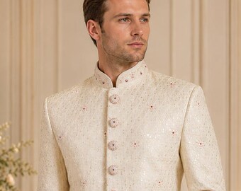 Può includere: Uno sherwani color bianco sporco con colletto alla coreana, maniche lunghe e abbottonatura frontale con bottoni rivestiti in tessuto coordinato. Il capo è decorato con un sottile motivo floreale ripetuto, accentuato da piccoli dettagli rossi.