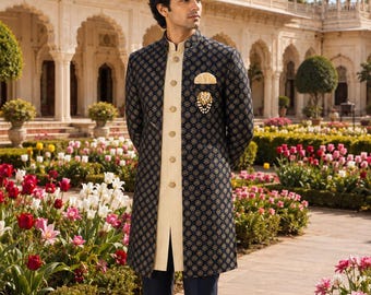May include: A navy blue sherwani featuring a repeating gold floral pattern, worn over a cream-colored button-down kurta and matching navy blue trousers. The outfit includes a gold pocket square and a decorative brooch with dangling pearls.