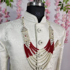 May include: A formal necklace with a silver-colored rectangular top and red and white beads. The necklace is layered and hangs over a white patterned jacket on a mannequin. The jacket has decorative buttons and a pocket square.