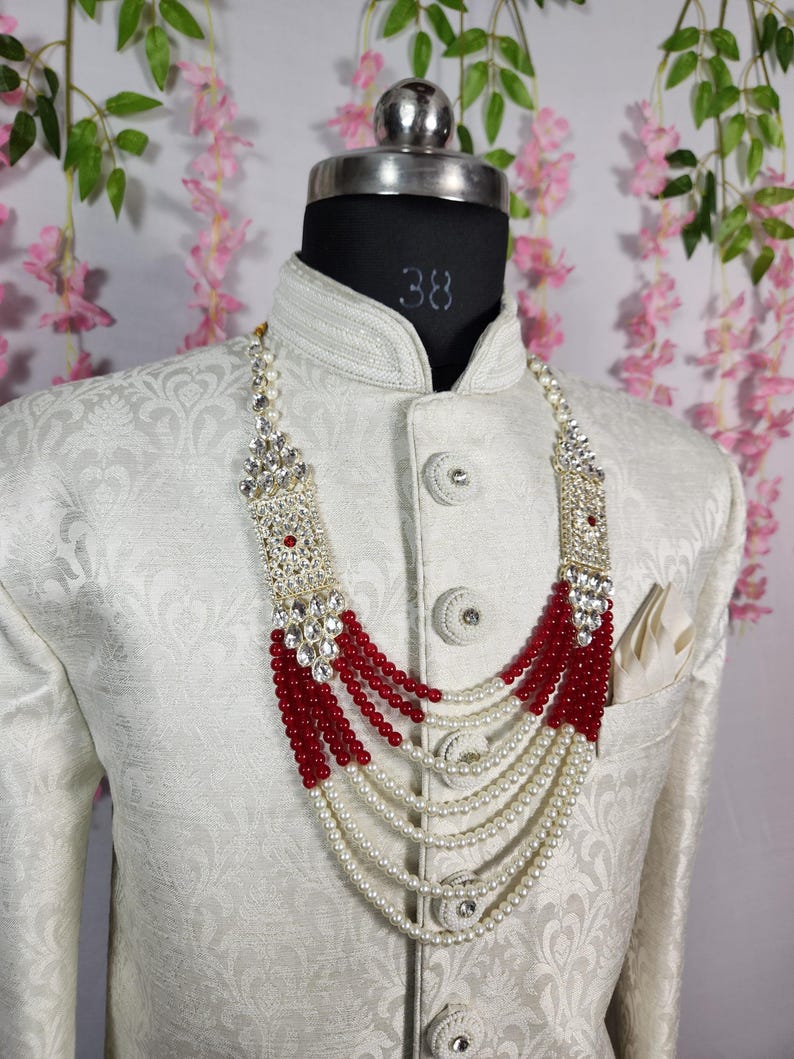 May include: A formal necklace with a silver-colored rectangular top and red and white beads. The necklace is layered and hangs over a white patterned jacket on a mannequin. The jacket has decorative buttons and a pocket square.