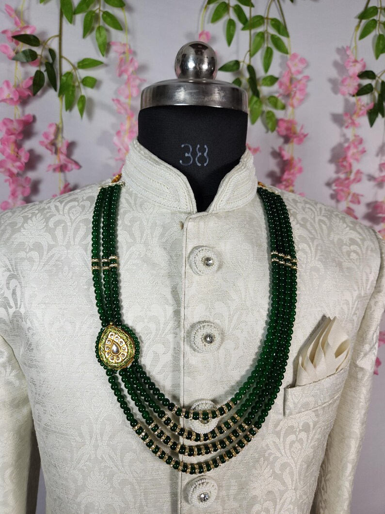 Puede incluir: Un collar de cuentas verdes de varias hebras con un colgante dorado con un centro en forma de perla. El collar se muestra sobre una chaqueta blanca estampada con un pa&ntilde;uelo de bolsillo. La chaqueta est&aacute; sobre una cabeza de maniqu&iacute; negra.