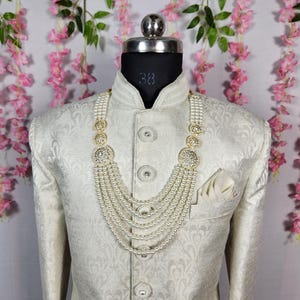 May include: A cream-colored, patterned jacket with a matching pocket square and a multi-strand pearl necklace with gold and crystal accents. The jacket is buttoned up and displayed on a mannequin.