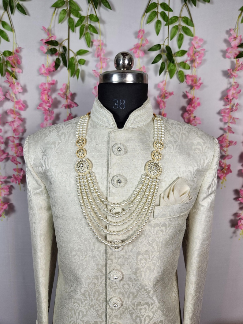 May include: A cream-colored, patterned jacket with a matching pocket square and a multi-strand pearl necklace with gold and crystal accents. The jacket is buttoned up and displayed on a mannequin.