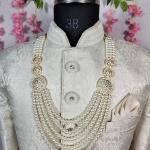 May include: A cream-colored, multi-strand pearl necklace with gold-toned, ornate accents. The necklace is displayed on a mannequin wearing a cream-colored, patterned jacket with a pocket square.