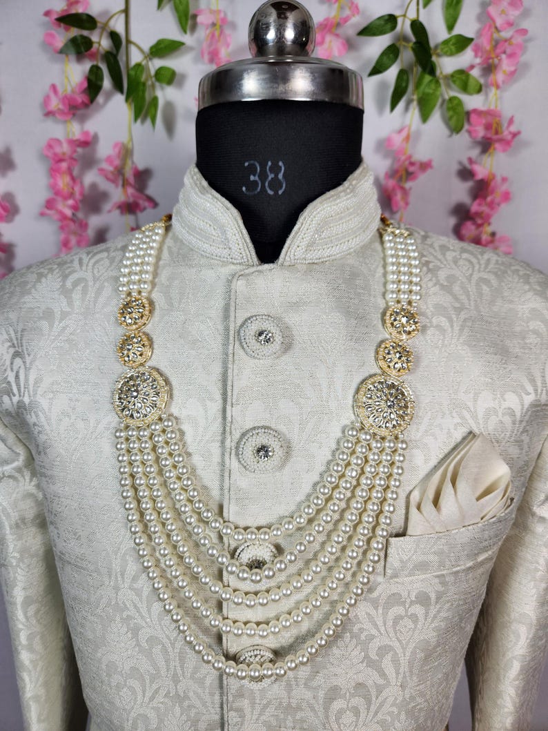 May include: A cream-colored, multi-strand pearl necklace with gold-toned, ornate accents. The necklace is displayed on a mannequin wearing a cream-colored, patterned jacket with a pocket square.