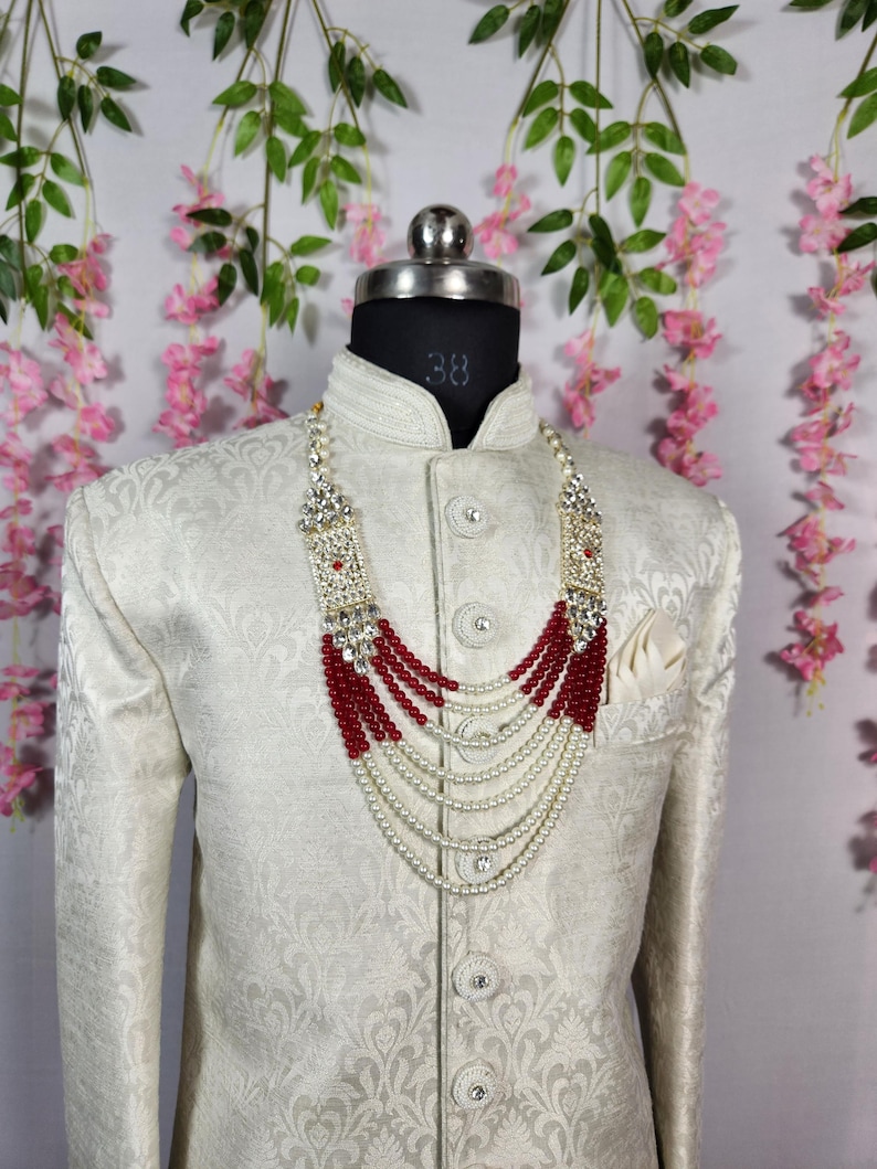 May include: A cream-colored, patterned jacket with a high collar and a decorative beaded necklace. The necklace features red and white beads, with a silver-colored clasp. The jacket is displayed on a mannequin.