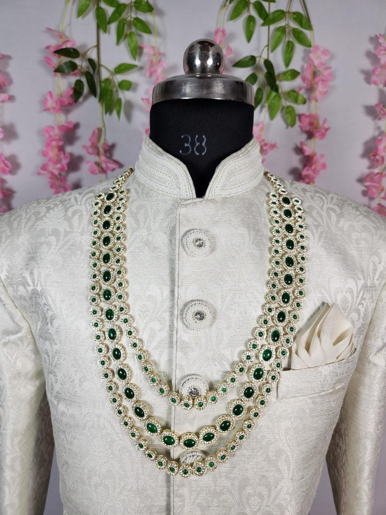 Pode incluir: Um colar de tr&ecirc;s fios com pedras verde esmeralda e detalhes brancos &eacute; exibido em um casaco branco. O casaco tem um padr&atilde;o texturizado e um len&ccedil;o de bolso. O colar &eacute; em camadas e ornamentado, com um design cl&aacute;ssico.