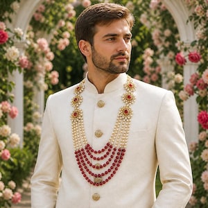 May include: A cream-colored suit with gold buttons and a mandarin collar. The suit is paired with a layered necklace of pearls and red beads. The background features a floral archway with pink and white roses.