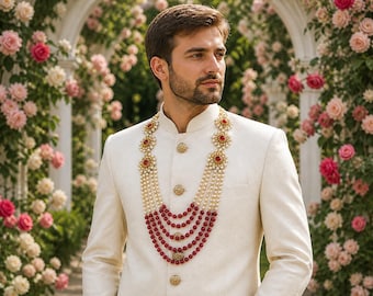 Pode incluir: Um terno creme com botões dourados e gola mandarim. O terno é combinado com um colar em camadas de pérolas e contas vermelhas. O fundo apresenta um arco floral com rosas rosa e brancas.