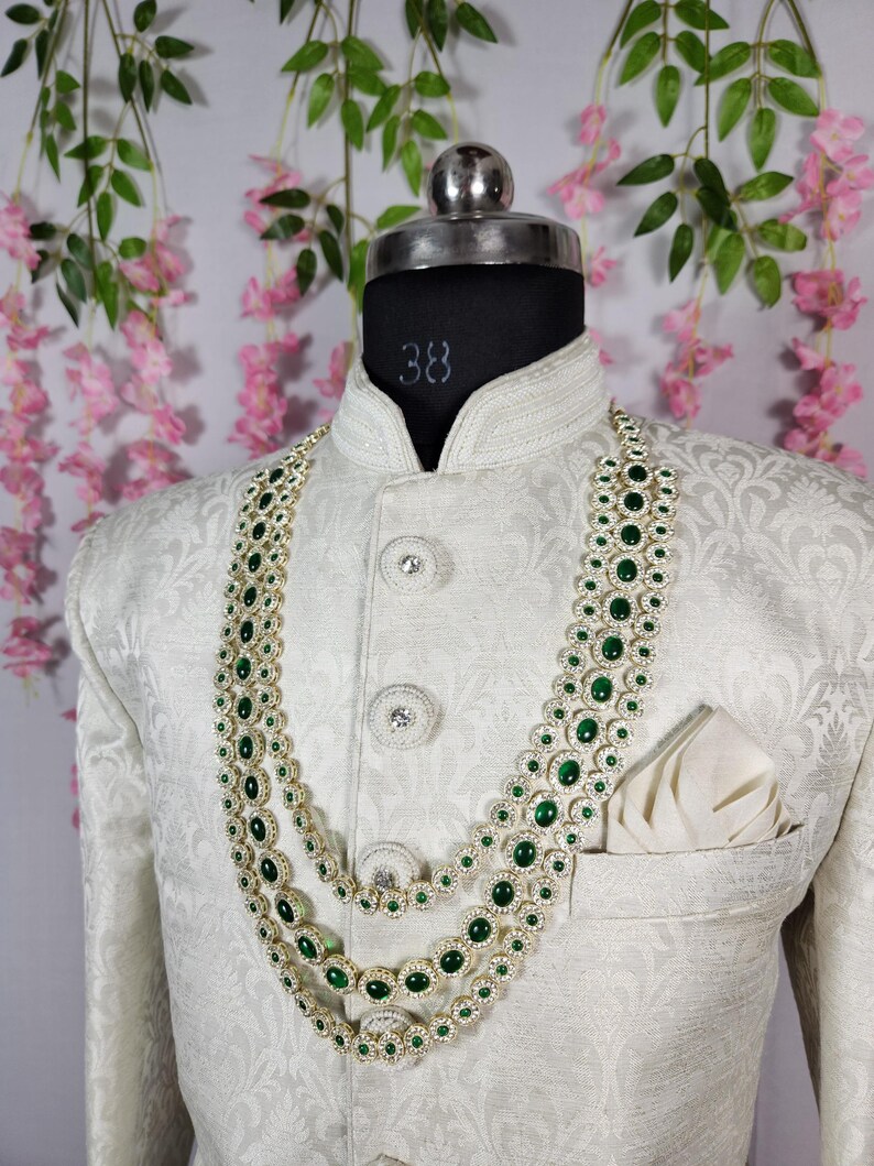 Pode incluir: Casaco creme com padr&atilde;o floral, usado por um manequim. O casaco &eacute; adornado com um colar de tr&ecirc;s fios com pedras preciosas verdes e uma moldura prateada. Um len&ccedil;o de bolso branco est&aacute; no bolso do peito.