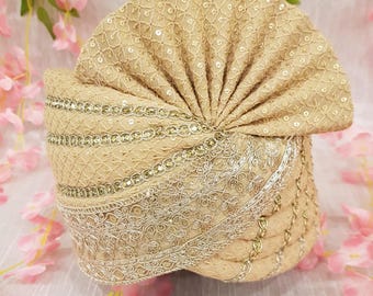 Pode incluir: Um turbante bege com bordados dourados intrincados e detalhes de lantejoulas. O turbante apresenta uma coroa em forma de leque e é adornado com delicadas rendas. Um adorno de cabeça tradicional.