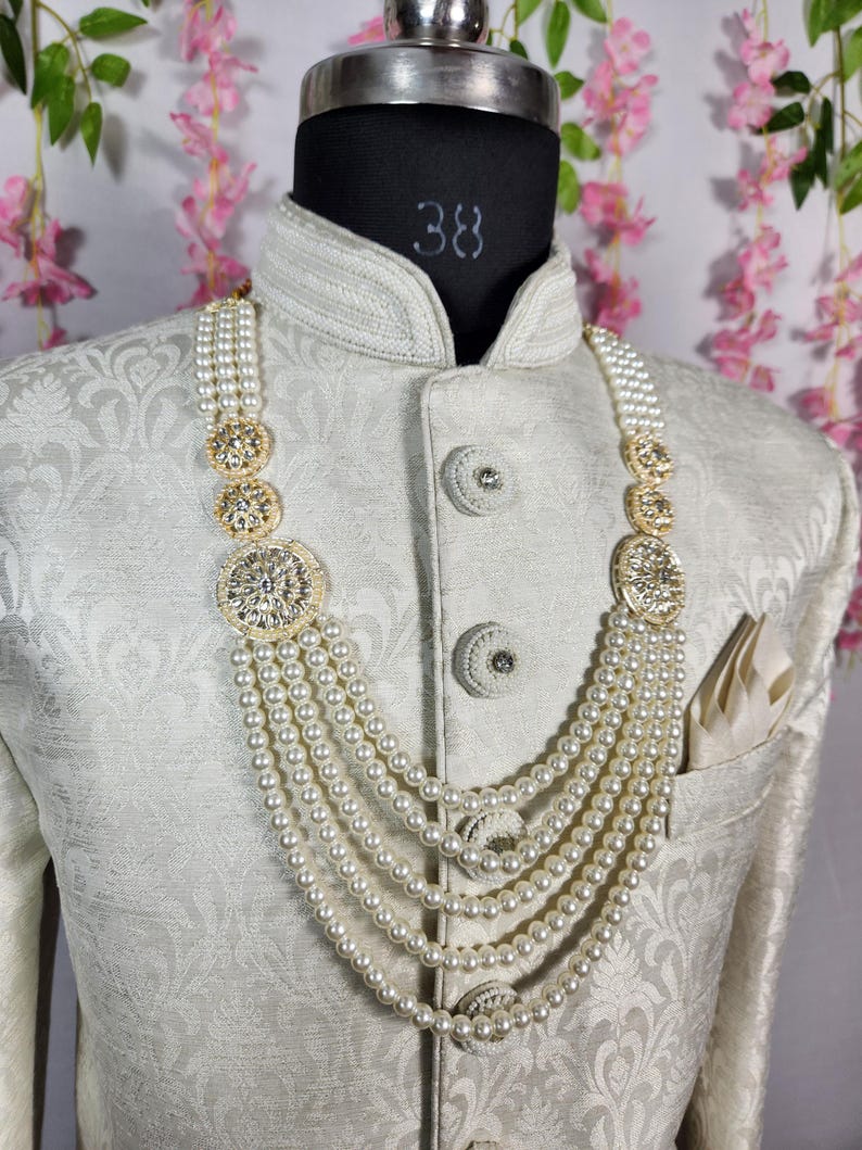 May include: A cream-colored, ornate necklace with multiple strands of faux pearls and gold-toned, circular embellishments. The necklace is displayed on a mannequin wearing a cream-colored, patterned jacket with a pocket square.