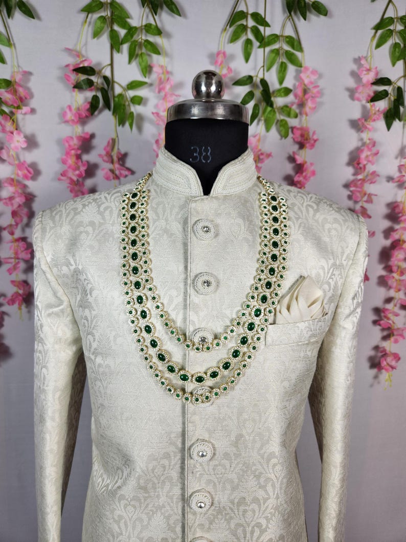 Pode incluir: Casaco creme estampado com gola mandarim e bot&otilde;es decorativos. Um colar de tr&ecirc;s fios com pedras preciosas verdes e detalhes prateados adorna o casaco. Um len&ccedil;o branco est&aacute; no bolso do peito. O casaco &eacute; exibido em um manequim.